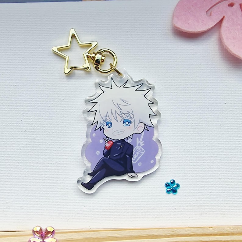 May include: A clear acrylic charm featuring a cartoon character with white hair and blue eyes, wearing a dark suit. The charm has a gold star-shaped clasp and a gold keyring. The character is sitting and holding a small red item and a drink.