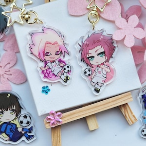 Könnte beinhalten: Fünf bunte Anime-Charakter-Schlüsselanhänger mit schwarzen Umrissen. Jeder Schlüsselanhänger zeigt einen anderen Charakter mit einem Fußball. Die Charaktere tragen verschiedene farbige Uniformen mit Nummern darauf. Die Schlüsselanhänger hängen an silbernen Ketten mit sternförmigen Anhängern.