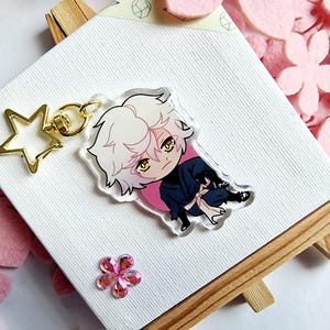 Könnte beinhalten: Ein klarer Acryl-Schlüsselanhänger mit einem Chibi-Anime-Charakter mit weißen Haaren, gelben Augen und einem dunkelblauen Outfit. Der Schlüsselanhänger hat einen goldfarbenen sternförmigen Verschluss und einen goldenen Schlüsselring. Der Charakter ist vor einem rosa Kreis platziert.