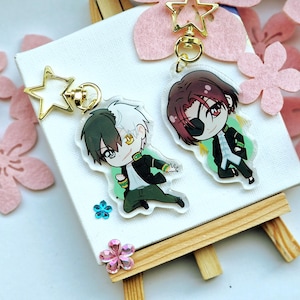 Könnte beinhalten: Zwei Anime-Charakter-Schlüsselanhänger mit goldenen Stern-Anhängern. Die Charaktere sind im Chibi-Stil gezeichnet. Ein Charakter hat weißes Haar und grüne Augen, der andere hat rotes Haar und ein Augenpflaster. Beide Charaktere tragen schwarze und grüne Outfits.