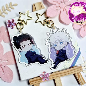 May include: Two anime-style acrylic keychains featuring characters in dark blue suits. One has black hair, the other white. Both are attached to gold star-shaped clasps. The background includes pink flower cutouts and the word "Chuuya".