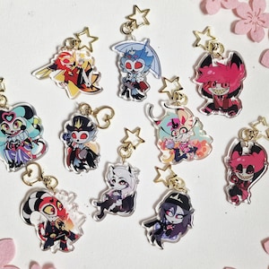 Könnte beinhalten: Eine Sammlung von Acryl-Anhängern mit bunten Cartoon-Charakteren. Jeder Anhänger hat einen goldfarbenen sternförmigen Schlüsselanhänger. Die Charaktere haben verschiedene Designs, darunter Dämonen und andere Fantasy-Figuren. Die Anhänger liegen auf einer weißen Oberfläche mit rosa Blumendekorationen.