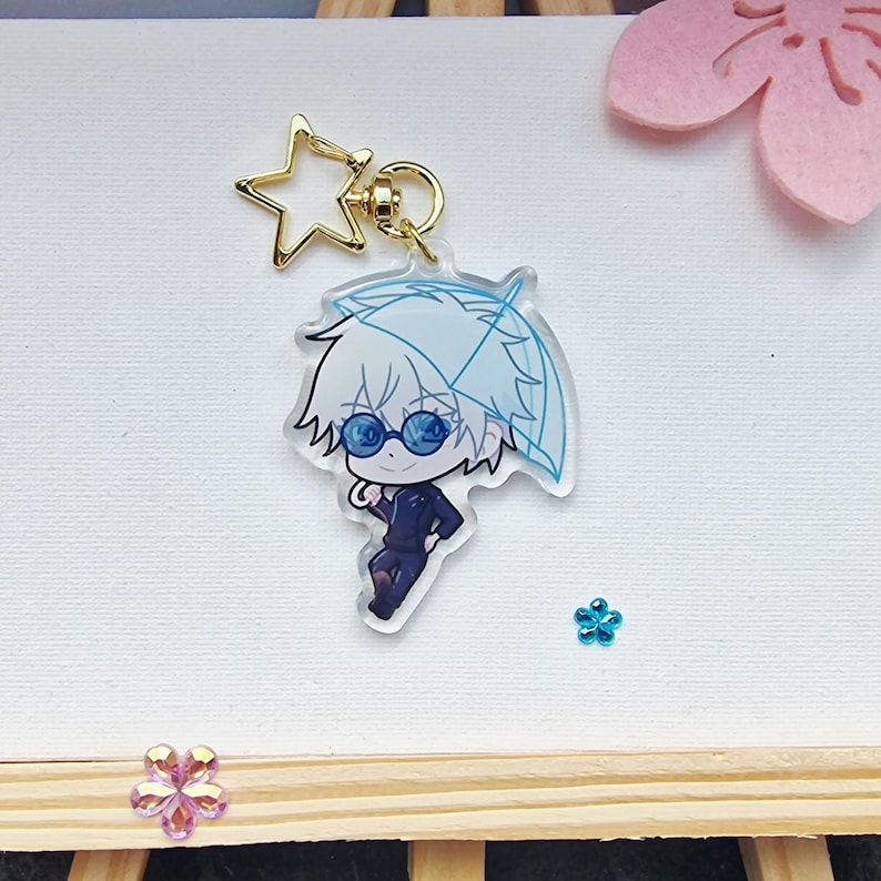 May include: A clear acrylic keychain featuring a cartoon character with white hair, blue sunglasses, and a blue umbrella. The keychain has a gold star-shaped charm and a gold ring. The character is wearing a dark blue outfit.