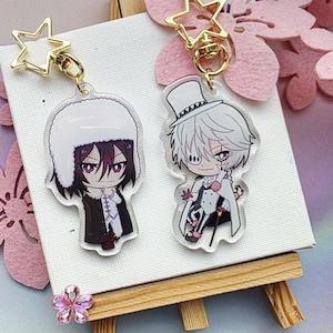Peut inclure: Deux porte-clés en acrylique représentant des personnages de style anime. L'un porte un chapeau de fourrure blanc et un manteau noir, l'autre un haut-de-forme et un manteau blanc. Les deux porte-clés ont des fermoirs dorés en forme d'étoile.