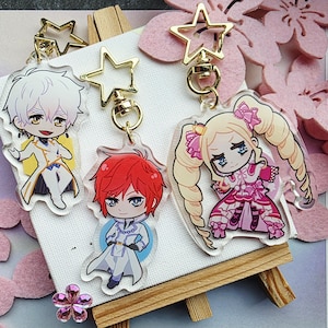 Op de afbeelding: Drie sleutelhangers met anime-personages. Elke sleutelhanger heeft een ander karakterontwerp, met goudkleurige stervormige sluitingen. De personages hebben verschillende haarkleuren en outfits. De sleutelhangers worden op een klein ezelbord gepresenteerd.