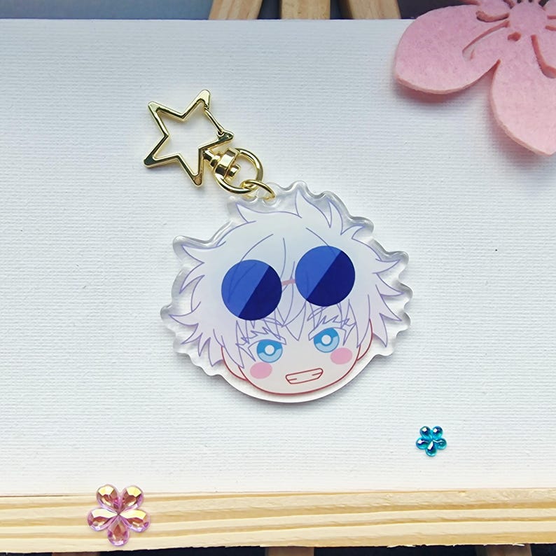 May include: A clear acrylic keychain featuring a cartoon character with white hair, blue eyes, and a smiling expression. The keychain has a gold star-shaped clasp and a gold ring. The character's face is detailed with pink cheeks and a small mouth.