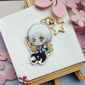 Könnte beinhalten: Ein klarer Acryl-Anhänger mit einem Anime-Charakter im Chibi-Stil mit weißen Haaren und einem schwarzen Outfit. Der Anhänger hat einen goldfarbenen sternförmigen Verschluss und einen goldenen Schlüsselring. Der Hintergrund umfasst rosa Blumendekorationen.
