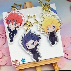 Könnte beinhalten: Drei Schlüsselanhänger aus Acryl mit Anime-Charakteren mit stacheligen Haaren und detaillierten Outfits. Jeder Schlüsselanhänger hat einen goldfarbenen sternförmigen Verschluss. Die Charaktere haben rote, gelbe und blaue Haare mit passenden Augen. Die Schlüsselanhänger werden auf einer kleinen Staffelei präsentiert.