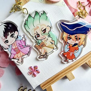 Könnte beinhalten: Drei Schlüsselanhänger im Anime-Stil aus Acryl mit farbenfrohen Charakteren und goldfarbenen Metallverschlüssen. Die Charaktere haben markante Frisuren und Outfits. Die Schlüsselanhänger werden auf einer weißen Oberfläche mit einer Holzstaffelei präsentiert.