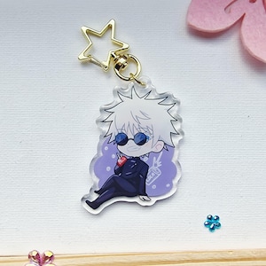 May include: A clear acrylic keychain featuring a cartoon character with white hair, sunglasses, and a black suit. The character is seated on a purple background and holding a red drink. The keychain has a gold star-shaped clasp.