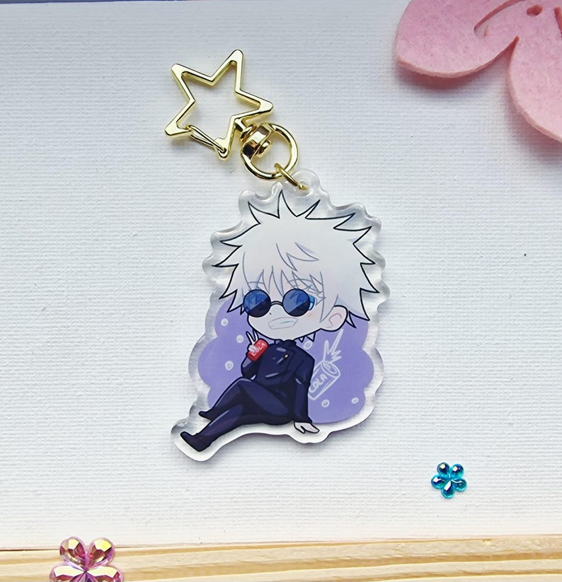 May include: A clear acrylic keychain featuring a cartoon character with white hair, sunglasses, and a black suit. The character is seated on a purple background and holding a red drink. The keychain has a gold star-shaped clasp.