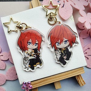 Könnte beinhalten: Zwei Schlüsselanhänger aus Acryl im Anime-Stil mit einer Figur mit roten Haaren und blauen Augen, jeweils mit einem goldfarbenen sternförmigen Verschluss. Die Figur ist mit einem kleinen weißen Tier dargestellt. Ein Schlüsselanhänger hat ein schwarz-weißes Outfit, der andere ein schwarz-goldenes Outfit.