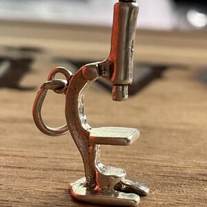 May include: A small, silver-colored microscope charm with a rounded base, a stage, and a vertical tube. The charm has a ring for attachment and is set against a wooden surface.