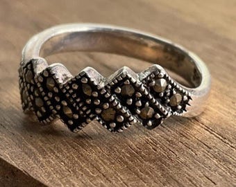 Puede incluir: Un anillo de plata con un patrón en zigzag de pequeñas piedras oscuras. El anillo se encuentra sobre una superficie de madera, mostrando su intrincado diseño y brillo metálico. La banda del anillo es lisa y pulida.