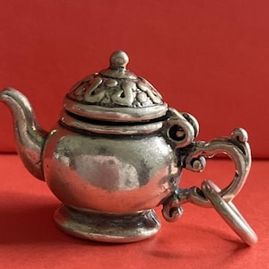 Może przedstawiać: Mały, srebrny charms w kształcie czajniczka z misternymi detalami na pokrywce i uchwycie. Czajniczek ma zaokrąglony korpus i mały dzióbek. Charms jest zawieszony na małym kółeczku. Tło jest jednolicie czerwone.