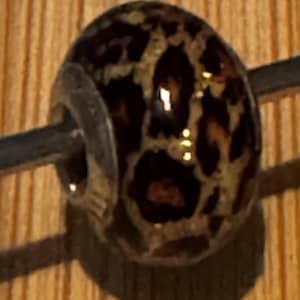 Puede incluir: Una cuenta redonda con un núcleo de metal plateado. La cuenta tiene un color base marrón oscuro con un estampado de leopardo en tonos beige y marrón. La cuenta está en una varilla de metal plateado.