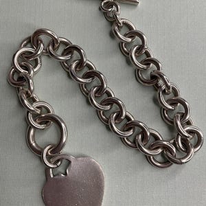 May include: A silver-toned chain link bracelet featuring a heart-shaped pendant attached to one end and a T-bar toggle closure at the other. The bracelet is constructed from a series of circular metal links connected in a continuous loop design.