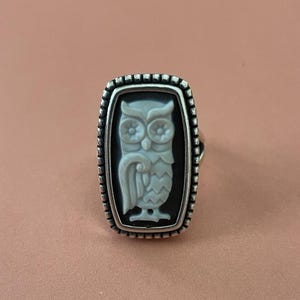 Op de afbeelding: Zilverkleurige ring met een rechthoekige zwarte camee met een wit uilontwerp. De uil is ingewikkeld gesneden en de ring heeft een kralenrand. Een uniek sieraad.
