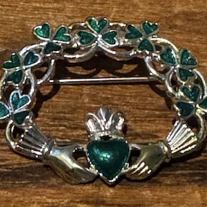 May include: A silver-toned brooch featuring a Claddagh design with a central green heart held by two hands, topped with a crown. The circular frame is decorated with a series of green enamel shamrock motifs arranged in a continuous wreath pattern.