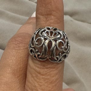 Puede incluir: Anillo de plata ornamentado con un diseño calado detallado. El anillo tiene una forma redondeada con patrones arremolinados y detalles florales. El metal tiene un acabado pulido, que refleja la luz. Una pieza de joyería llamativa.