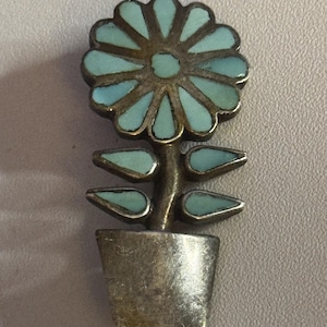 May include: A silver-toned flower pin with a turquoise blue flower and leaves, set in a pot. The flower has a detailed design with individual petals. The pin is likely made of metal and is a decorative accessory.
