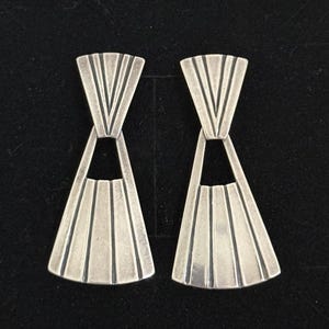May include: A pair of silver-toned earrings with a geometric design. Each earring features a fan-shaped bottom with vertical lines and a V-shaped top. The earrings are connected by a small, open section.