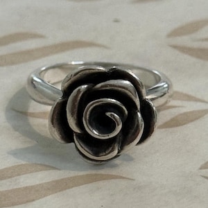 Puede incluir: Un anillo de plata con un diseño de rosa detallado. La rosa es el punto focal, con intrincados detalles de pétalos. La banda del anillo es lisa y pulida, creando un contraste con la rosa texturizada. El anillo es una joya.
