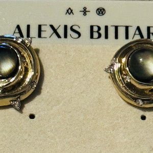 Puede incluir: Un par de pendientes dorados con un diseño en espiral. Cada pendiente presenta una piedra central oscura e iridiscente y está adornado con pequeños cristales transparentes. La marca "ALEXIS BITTAR" es visible encima.