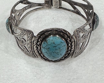 Op de afbeelding: Zilverkleurige scharnierende armband met turkooizen stenen. De armband heeft een ovale turkooizen steen in het midden, omgeven door een gevlochten ontwerp. De zijkanten hebben geëtste bloempatronen en kleinere turkooizen stenen. De armband is ongeveer 5 cm breed.