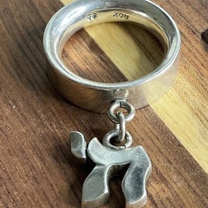 May include: A silver ring with a thick band and a dangling pendant. The pendant is in the shape of the Hebrew letter "Chai". The ring is on a wooden surface.