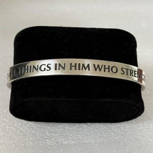 May include: A silver-colored cuff bracelet with the engraved text "ALL THINGS IN HIM WHO STRENGTH". The bracelet is displayed on a black velvet surface. The bracelet is a simple, elegant design.