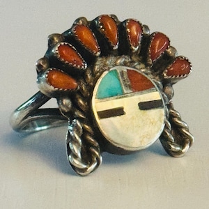 May include: A silver ring featuring a detailed design. The ring has a central circular element with turquoise, red, and white inlays, and a black horizontal bar. It is surrounded by orange teardrop-shaped stones and decorative silverwork.