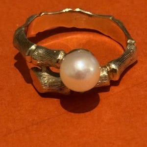 May include: A gold-colored ring with a bamboo-style band and a large, round, white pearl. The ring is set against an orange background, highlighting the contrast between the warm gold and the cool pearl.