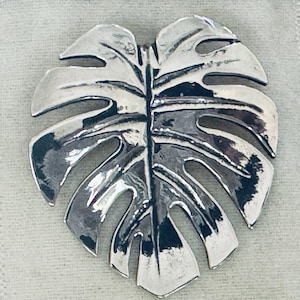 May include: A silver-toned metal leaf-shaped brooch. The brooch is a detailed representation of a Monstera plant leaf, with cut-out sections and a central vein. The surface has a polished, reflective quality, and the overall design is botanical.