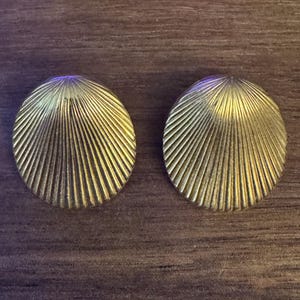 May include: Two gold-colored, round, shell-shaped earrings. Each earring has a ridged, fan-like design radiating from the center. The earrings are displayed on a dark brown wooden surface.