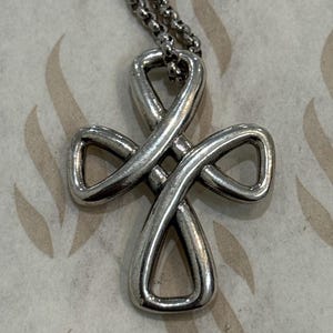 Puede incluir: Un collar con colgante plateado con un diseño de cruz abierta. La cruz está formada por metal en bucle, creando una forma de símbolo de infinito. La cadena es de diseño de eslabones. El fondo es de color beige claro con un patrón de hojas.