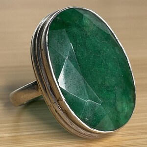 May include: A silver ring featuring a large, oval-shaped, faceted green gemstone set in a smooth metal bezel. The stone displays deep emerald tones with visible internal inclusions and a polished surface, held by a simple, solid silver band.