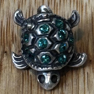 Puede incluir: Figura de tortuga plateada con caparazón texturizado adornado con piedras preciosas verdes. La tortuga tiene aletas, cabeza y cola detalladas, sobre un fondo de madera.