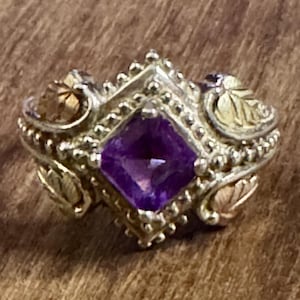 May include: A silver ring with a square, deep purple gemstone at the center. The ring features gold-toned leaf accents and beaded detailing around the gemstone. The ring is set on a wooden surface.