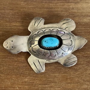 May include: A silver-toned turtle-shaped pendant with a turquoise stone center. The pendant has detailed etchings around the stone and on the turtle's shell and flippers. The pendant is displayed on a wooden surface.