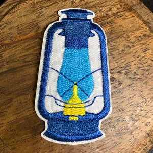 May include: Embroidered patch featuring a vintage lantern design. The patch has a white background with a blue outline and details. The lantern's glass is a gradient of blue, and the base is yellow. The patch is likely for clothing or accessories.