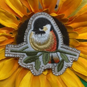 May include: An embroidered patch featuring a small bird with a black head, white cheeks, and a golden-brown chest perched on a green branch. The patch is outlined with a border of small, clear, reflective crystals on a light gray felt backing.