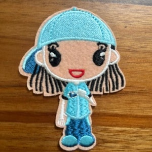 May include: Embroidered iron-on patch of a cartoon girl with black hair, wearing a blue baseball cap, blue shirt, and blue jeans.