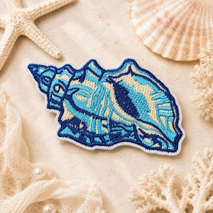 May include: An embroidered patch in the shape of a conch shell featuring a color palette of dark blue, bright turquoise, and cream. The patch has a white border and intricate thread work detailing the spiral structure of the seashell.
