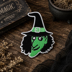 May include: An embroidered patch featuring a green-skinned witch with black hair and a black pointed hat. The hat has a light green band with a white buckle. The entire patch is outlined in white thread, showcasing a detailed stitched design.