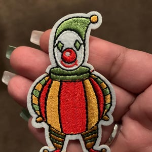 May include: Embroidered clown patch with a white face, green hat, and a red nose. The clown's body is a combination of red, yellow, and green stripes. The patch is outlined in black and has a white border.