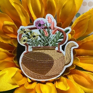 May include: An embroidered patch shaped like a brown teapot filled with a bouquet of yellow, purple, and pink flowers. The teapot has a curved handle and spout, with detailed stitching in shades of brown and tan, set against a white border.