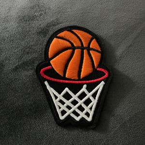 May include: Embroidered patch featuring a basketball going through a hoop. The basketball is orange with black lines, and the hoop is black with a red rim and white net. This patch is suitable for sports enthusiasts.