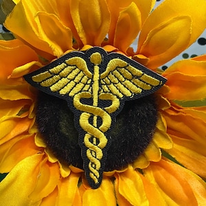May include: A black embroidered patch featuring a gold caduceus symbol with two wings and two snakes coiled around a central staff. The patch is positioned against the center of a large, vibrant yellow sunflower with dark brown petals.