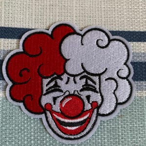 May include: Embroidered patch featuring a smiling clown face with red and white curly hair, a red nose, and a wide red and black smile. The patch has a white background and a black border.